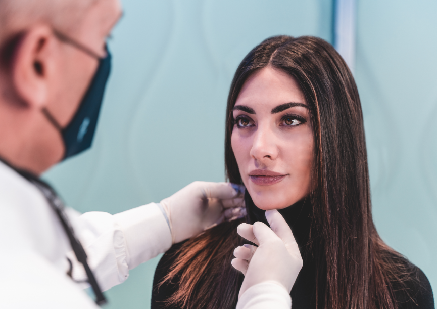 A practitioner wearing a face mask and white gloves examining a female client's jawline during a consultation.