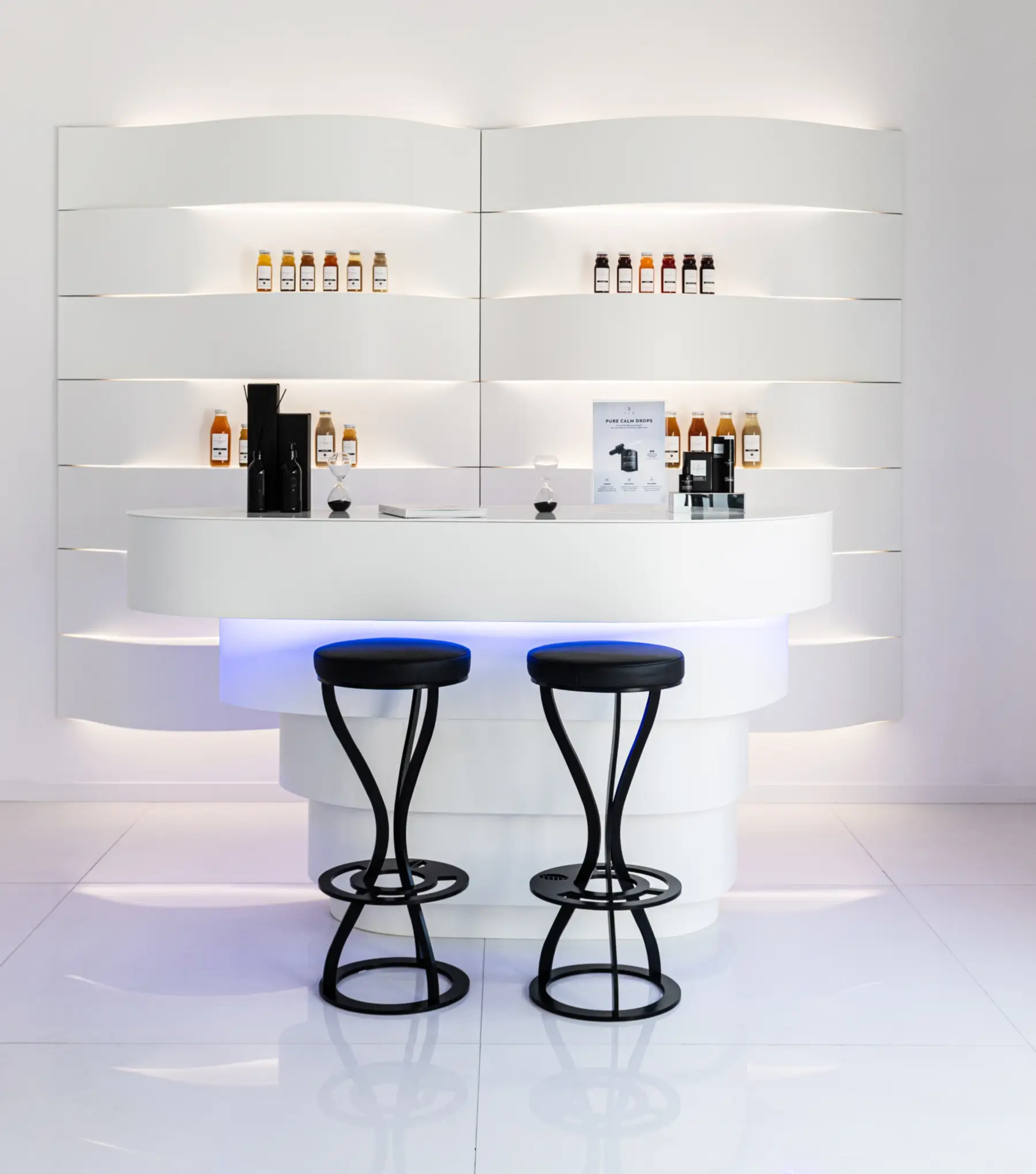 Modern, minimalist white health and wellness bar featuring lighted shelves with stacked supplement bottles, a sleek white counter, and two tall black wireframe barstools.