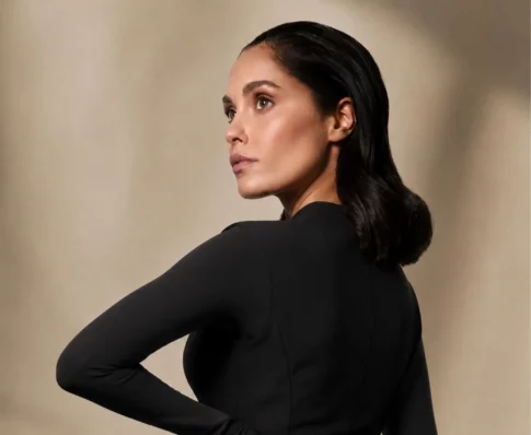 A woman with slicked-back dark hair turned in profile, wearing a black top against a warm beige background.
