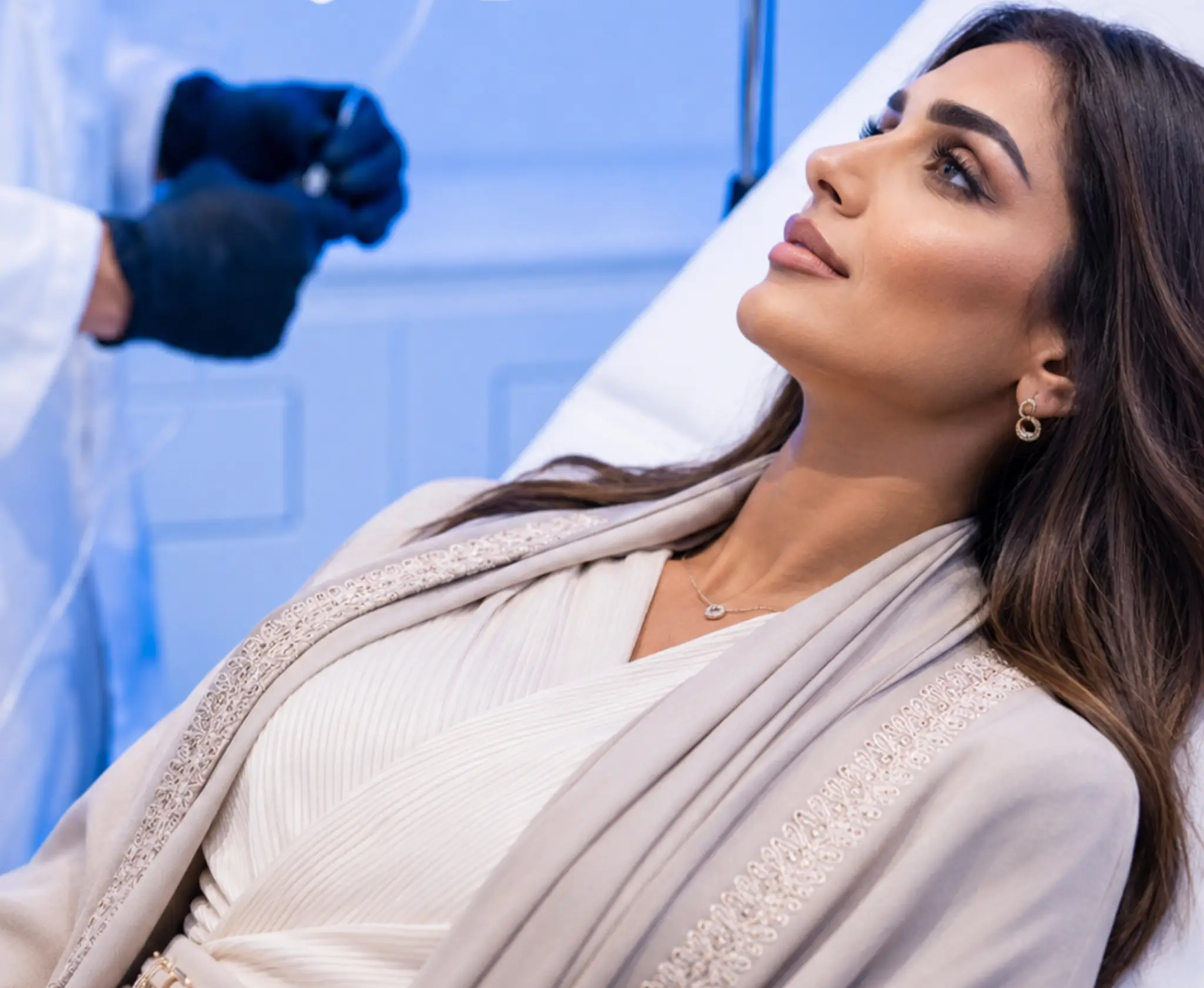 A woman sitting in a clinic treatment chair receiving IV drip therapy or an injectable procedure from a medical professional wearing black gloves.