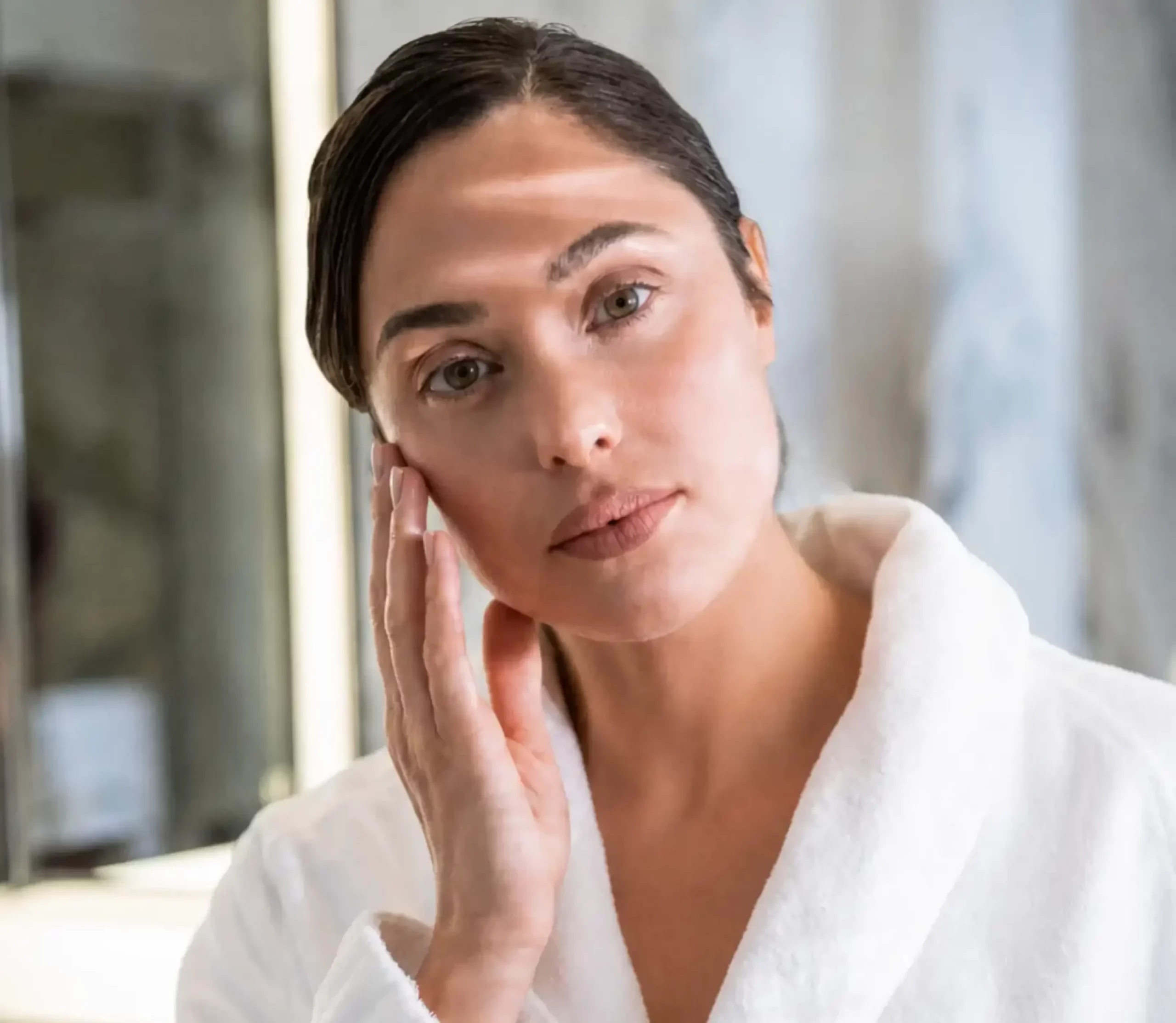 A woman in a white bathrobe resting her hand on her cheek while looking into the camera.