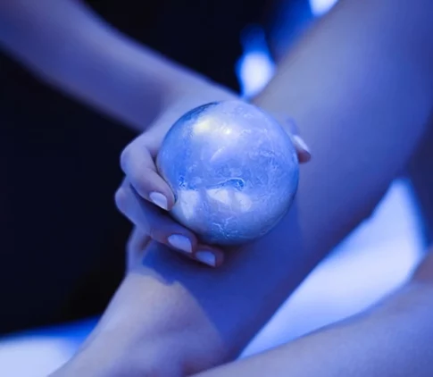 A hand holding an ice sphere against a leg under cool blue lighting.