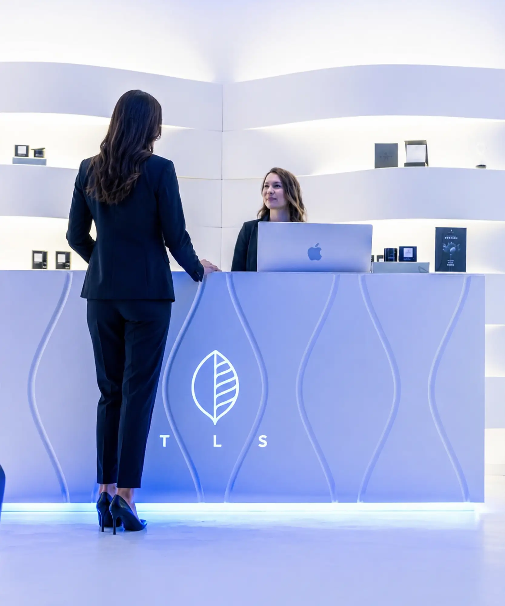 A customer standing at a modern, backlit white reception desk with the TLS leaf logo, speaking with a receptionist.
