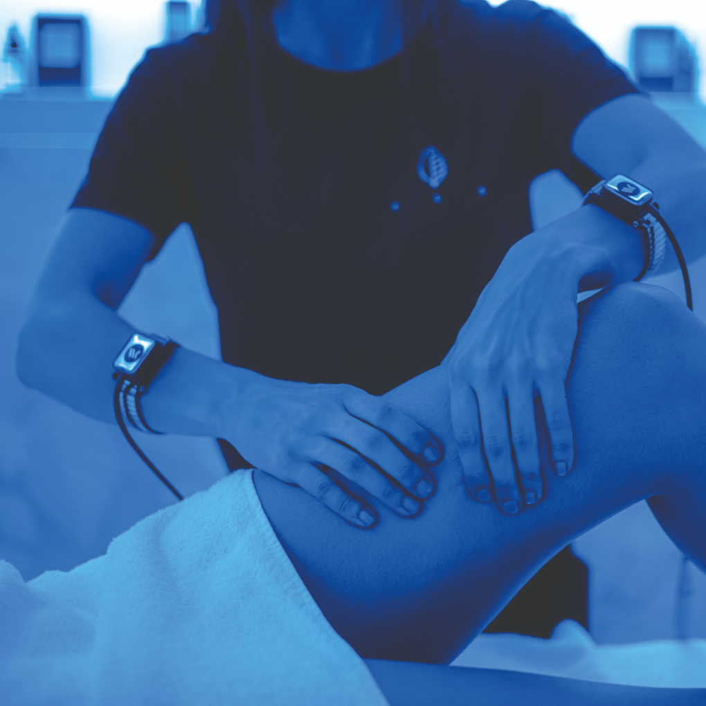 A practitioner wearing specialized wearable devices performs a manual leg massage treatment on a client in a cryotherapy or wellness setting illuminated by blue light.