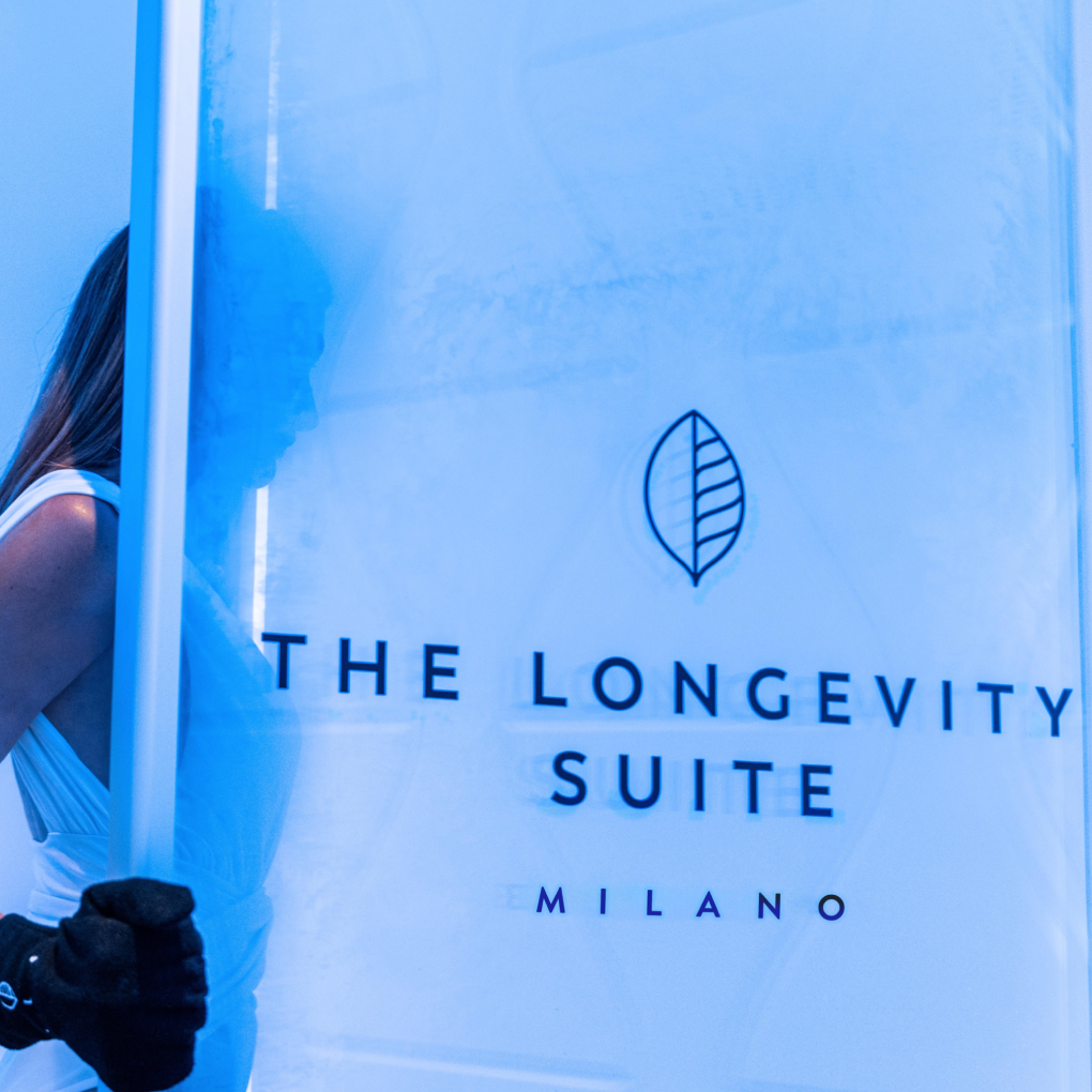 A woman inside a blue-lit cryotherapy chamber featuring 'The Longevity Suite Milano' logo on the clear glass door.