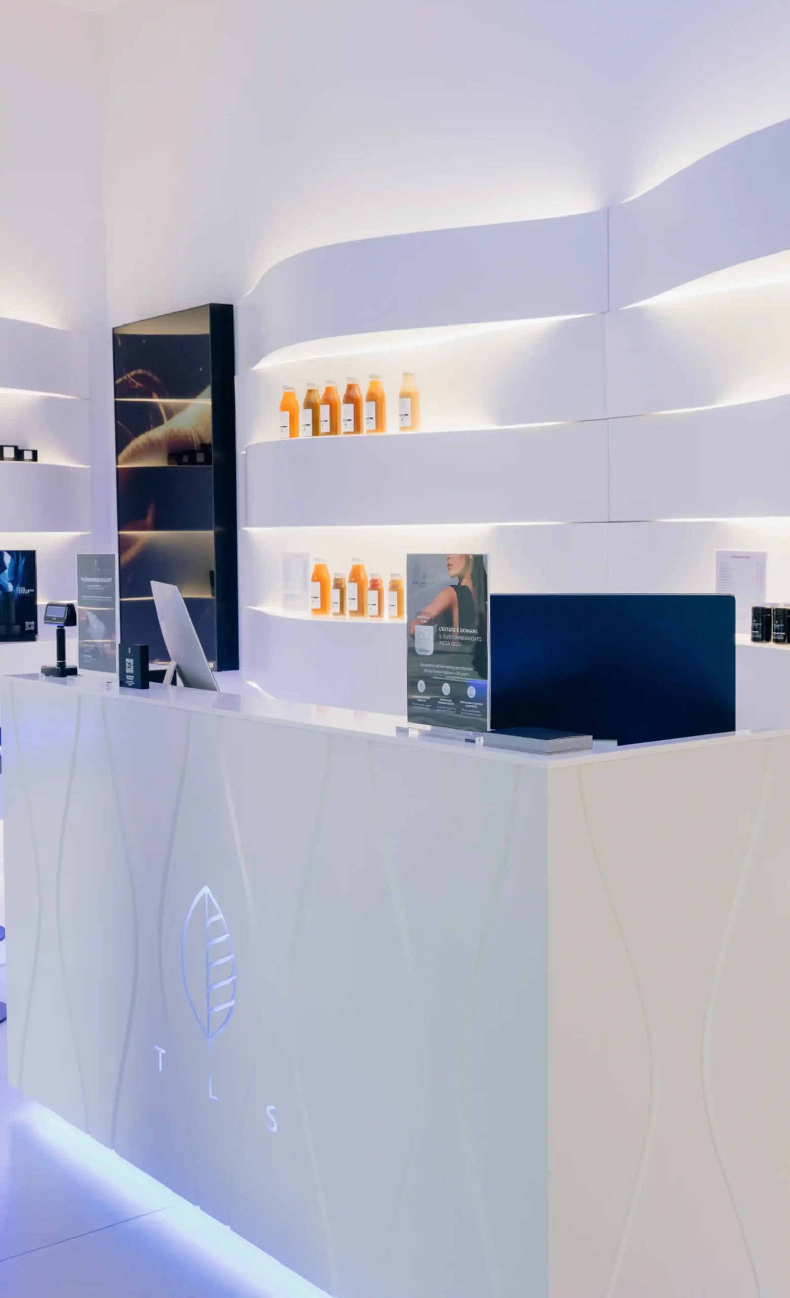 Modern white reception desk at The Longevity Suite with an illuminated TLS logo and backlit curved shelving displaying wellness products.