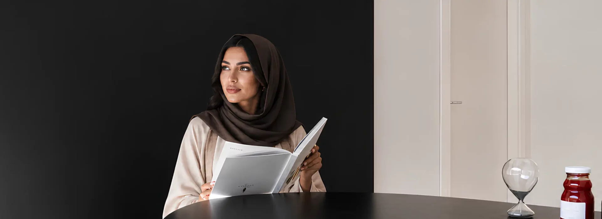 A woman wearing a brown hijab and light abaya reading a brochure at a dark table, with a modern glass hourglass and jar nearby.