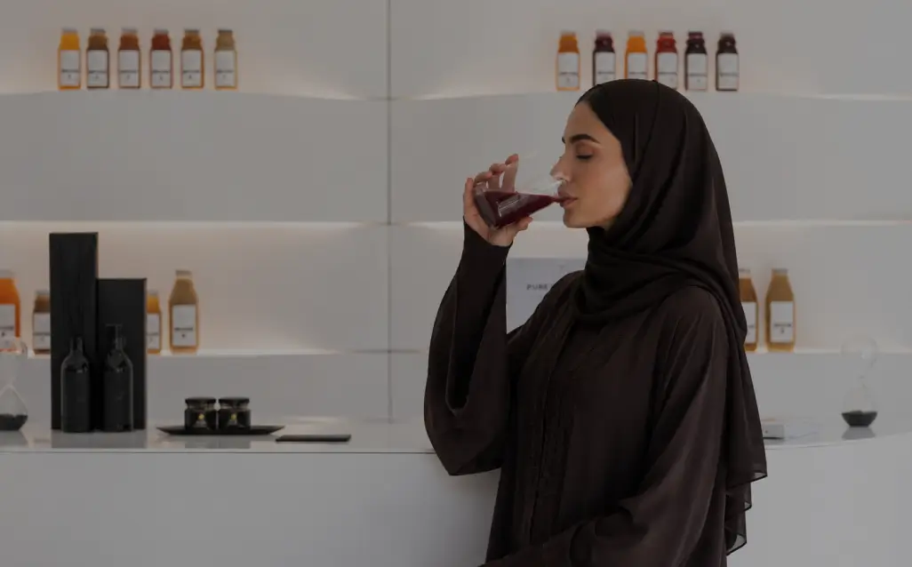 A woman wearing a black abaya and hijab drinking a dark beverage from a glass inside a modern, brightly lit wellness suite with skincare and supplement bottles displayed on white shelves in the background.