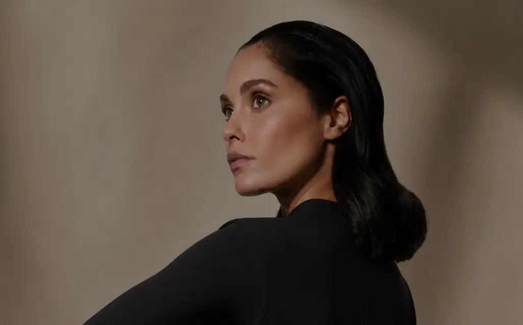 A woman with dark hair styled in loose waves and radiant skin, wearing a black long-sleeved top against a soft neutral background.
